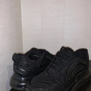 Svarta Nike Air Max 720 sneakers - Nike Air Max 720 sneakers i helsvart färg med mesh-överdel och synlig Air-sula som sträcker sig runt hela skon. Snygga vågformade detaljer och diskret Swoosh på sidan. Skorna har snörning och en rund tå, perfekt för dig som gillar sportig och modern stil.