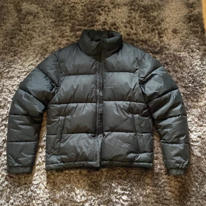 Svart pufferjacka från Everest - Svart pufferjacka från Everest med hög krage och dragkedja framtill. Jackan har en quiltad design och är tillverkad i syntetmaterial, perfekt för kalla dagar. Klassisk och stilren look som funkar till det mesta. Snygg passform och långärmad modell.