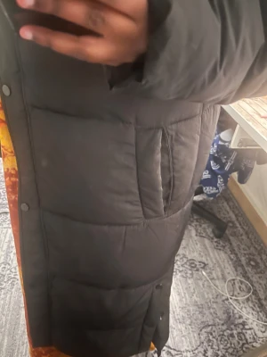 Svart lång pufferjacka med luva - Säljer en svart lång pufferjacka från Nelly med luva och tryckknappar med dragkedja längs hela framsidan. Jackan har quiltad design och är perfekt för kalla dagar. Materialet är syntet och jackan har långa ärmar. Enkel och stilren look som funkar till det mesta.