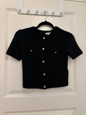 Svart kofta med guldfärgade knappar Gina Tricot - Svart, kortärmad kofta från Gina Tricot med snygga guldfärgade knappar framtill och på de två bröstfickorna. Koftan är stickad och har ribbade kanter vid hals, ärmslut och nederkant. Perfekt för dig som gillar klassisk och stilren look.