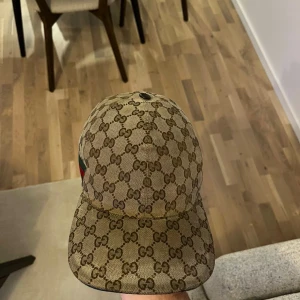 Gucci keps med GG-mönster - Snygg beige keps från Gucci med klassiskt GG-monogram över hela kepsen. Har justerbar rem bak och en grön-röd rand på sidan. Helt äkta ny pris 6,500. Materialet är slitstark bomullscanvas och kepsen har en böjd skärm. Perfekt för dig som vill ha en lyxig streetstyle-look.