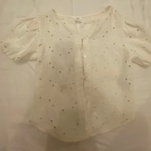 Vit blus med puffärm från Zara - Superfin vit blus från Zara med korta puffärmar och broderade hålmönster över hela tyget. Blusen har knappar framtill och är gjord i ett lätt, luftigt material som passar perfekt till varmare dagar.
