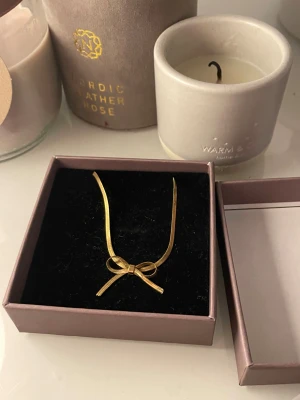 Guldigt halsband med rosett - Snyggt guldigt halsband med tunn kedja och en söt rosettdetalj framtill. Perfekt accessoar för att lyfta din outfit med en feminin touch. Justerbar längd med kedjeförlängning och smidig karbinhake. Kommer i en fin ask.
