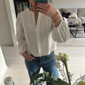 Vit broderad blus från Zara - Superfin vit blus från Zara med broderade detaljer och v-ringning. Blusen har ballongärmar med elastiska muddar och knappar framtill. Tillverkad i bomull och har ett romantiskt, luftigt uttryck som känns både trendigt och klassiskt.