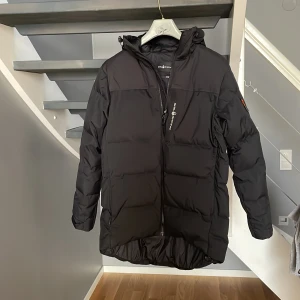 Svart Sail Racing dunjacka S - Svart dunjacka från Sail Racing med huva och snygga detaljer som Sail Antarctica Expedition-märke på ärmen. Jackan har dragkedja framtill, flera fickor och är designad för att hålla dig varm under kalla dagar. Perfekt för dig som gillar stilren och funktionell vinterjacka.