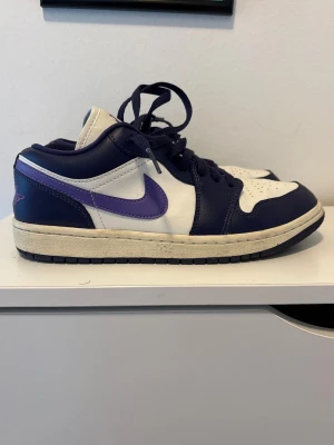 Nike Air Jordan 1 Low vit/lila - Original Nike Air Jordan 1 Low sneakers i vit och mörklila med lila detaljer och broderad Air Jordan-logga på hälen och Jumpman-logga på tungan. Har använd mindre än 5 gånger. Nästan helt nya! Kan skicka fler bilder i dm. Storlek EUR 40 (25,5 cm)  men passar 39-40. Priset kan diskuteras.