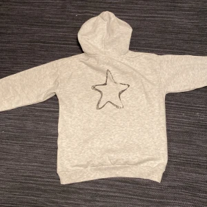 Grå hoodie från Gina Tricot - Snygg ljusgrå hoodie från Gina Tricot med huva och snörning. Framsidan har ett stort svart tryck med texten 'AMOUR' och baksidan en stjärna i svart. Mjuk insida och ribbade muddar vid ärmslut och nederdel.