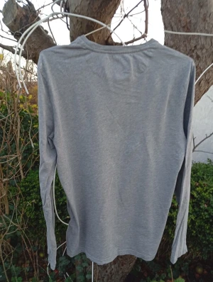 Grå långärmad tröja från Lyle & Scott - Snygg grå långärmad tröja från Lyle & Scott med klassisk rund hals och ikonisk gul broderad fågel på bröstet. Tillverkad i mjuk bomull, perfekt för lager-på-lager och chill vardagsstil.