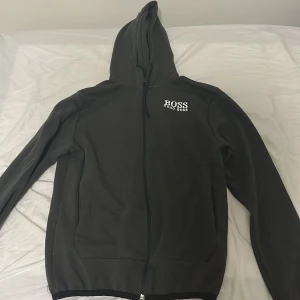 Svart hoodie från Hugo Boss - Olivgrön hoodie från Hugo Boss med vit broderad logga på bröstet. Tröjan har huva med snörning, hel dragkedja framtill och två sidofickor. Materialet är mjuk bomull och modellen är normal passform med långa ärmar. Perfekt för chill dagar eller när du vill ha en clean look.