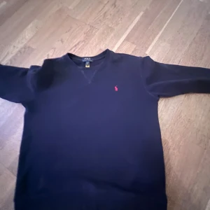 Mörkblå tröja från Polo Ralph Lauren - Snygg mörkblå tröja från Polo Ralph Lauren med klassisk röd broderad logga på bröstet. Tröjan har rund hals och långa ärmar, tillverkad i mjuk bomull. Perfekt för dig som gillar stilrena och tidlösa plagg med en stilren touch.