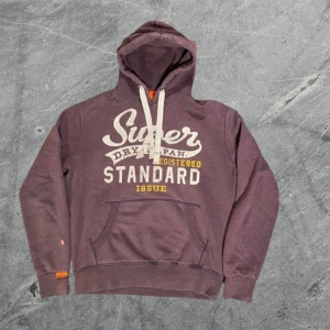 Superdry Hoodie - Säljer en lila hoodie från Superdry med stort vintage-tryck i vitt och gult på bröstet. Tröjan har huva med breda snören, magficka och ribbade muddar. Materialet är mjuk bomull. Säljer varan i befintligt skick. Passar bra till det mesta. Om du undrar något är det bara att skriva🙌✅