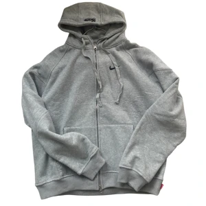 Grå hoodie Nike x Supreme Air Max - Grå hoodie från Nike och Supreme Air Max-kollektionen med stor svart swoosh på ryggen och mindre logga på bröstet. Tröjan har huva med snörning, dragkedja framtill och ribbade muddar. Tillverkad i mjuk bomull och polyesterblandning. Röd Supreme-tag på sidan.