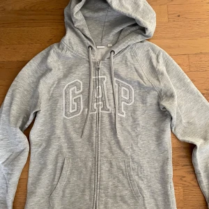 Grå hoodie från GAP med dragkedja - Grå hoodie från GAP i storlek S. Tröjan har broderad logga framtill, snörning vid huvan och dragkedja. Tillverkad i mjuk bomullsmix med långa ärmar och fickor framtill. Bra skick men en liten defekt på ena snöret då själva kanten gått upp. 