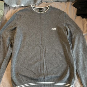 Grå långärmad tröja från Hugo Boss - Snygg grå tröja från Hugo Boss med diskret logga på bröstet. Rund hals med vit kant och ribbade muddar med vita ränder vid ärmslut och nederkant. Perfekt för dig som gillar stilrena och klassiska plagg.