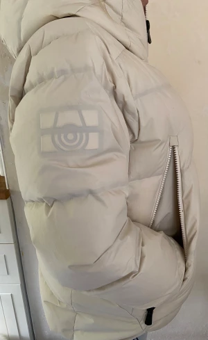 Beige pufferjacka från Sail Racing - Säljer en khakifärgad dunjacka från Sail Racing med hög krage luva och dragkedja framtill. Jackan har två sidofickor med dragkedja och ett stilrent Sail Racing-logomärke på ärmen. Materialet är dunfyllt, vindtätt och andas, perfekt för kalla dagar.