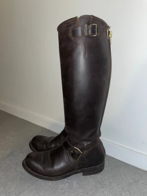 Primeboots bruna höga läderstövlar - Snygga höga stövlar från Primeboots i mörkbrunt skinn med mässingsfärgade spännen och dragkedja baktill. Klassisk rund tå och platt sula, perfekt för dig som gillar stilren och robust look. Passar till både jeans och kjol för en cool vibe.