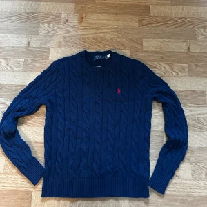 Marinblå stickad tröja Polo Ralph Lauren - Snygg marinblå stickad tröja från Polo Ralph Lauren med klassiskt kabelstickat mönster och röd broderad logga på bröstet. Rund halsringning och ribbade muddar vid ärmslut och nederkant. Perfekt för dig som gillar stilrena och tidlösa plagg. Helt ny, priset är ej hugget i sten så var inte rädda att ställa frågor 😁