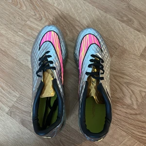 Nike Hypervenom silver fotbollsskor - Nike Hypervenom fotbollsskor i silver med neon-gul sula och insida, svart snörning och detaljer i rosa och guld. Skorna har ett mönstrat yttre med NikeSkin-material och en döskalle-detalj på hälen. Perfekt för dig som vill sticka ut på planen. Helt nya inta använda, priset kan diskuteras så var inte rädda att ställa frågor 😁
