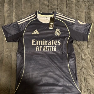 Real Madrid marinblå matchtröja Adidas S - Säljer en officiell Real Madrid fotbollströja från Adidas i marinblått med tunna ljusa ränder och vita detaljer. Tröjan har korta ärmar, rund hals och broderat klubbmärke samt Adidas-logga. Materialet är lätt polyester med Aeroready-teknologi.