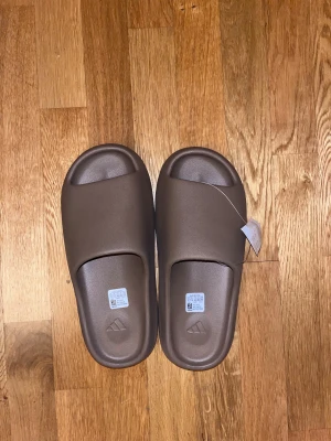 Adidas bruna slides med räfflad sula - Säljer ett par bruna Adidas slides med chunky, räfflad sula och stilren design. De har en bred rem över foten och är tillverkade i syntetmaterial för maximal komfort. Perfekta för chill dagar eller att glida runt hemma. Adidas-loggan är diskret präglad på fotbädden.