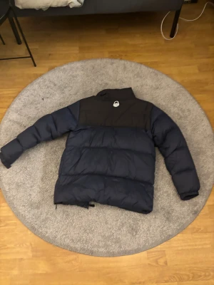 Mörkblå pufferjacka från Sailracing - Säljer en mörkblå pufferjacka från Sailracing med svart axelparti och hög krage. Jackan har långärmade ärmar, dragkedja framtill och ett stilrent, sportigt utseende. Perfekt för kalla dagar och har en modern, loose passform.