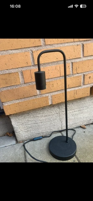 Svart bordslampa med modern design - Stilren svart bordslampa med minimalistisk design. Passar perfekt som skrivbords- eller läslampa. Lampan har en rund fot, böjd arm och cylinderformad lampskärm. Använd i 2 månader säljs pga flytt. Strömbrytare på sladden.