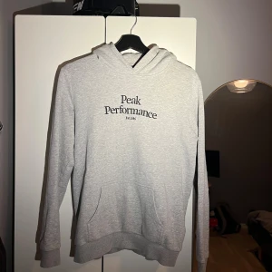 Grå hoodie från Peak Performance - Säljer en stilren grå hoodie från Peak Performance med svart broderad logga på bröstet. Tröjan har huva, magficka och ribbade muddar vid ärmslut och nederkant. Perfekt för chill dagar och enkel att matcha med jeans eller joggers.