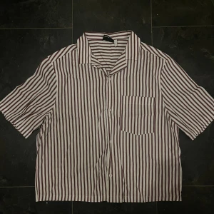 Randig kortärmad skjorta i vinrött - Snygg skjorta med vertikala vinröda och vita ränder, klassisk krage och bröstficka. Kortärmad och med knappar framtill, perfekt för dig som gillar en chill och stilren look. Materialet känns lätt och luftigt, passar bra till varma dagar.