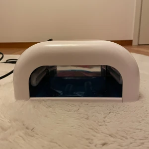 UV Nagellampa - Effektiv UV-lampa för härdning av gelnaglar. Vit plastkonstruktion med blå insida och ventilationshål på ovansidan. Lampan är i gott skick utan synligt slitage eller defekter. Perfekt för både hemmabruk och salong. Strömkabel medföljer.