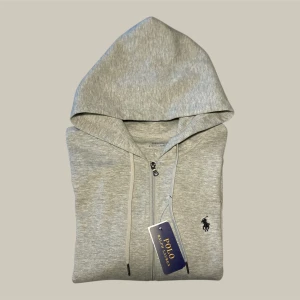 Grå hoodie från Polo Ralph Lauren - Snygg ljusgrå hoodie från Polo Ralph Lauren med dragkedja, huva och snörning. Klassisk broderad logga på bröstet och mjukt material i bomull. Perfekt för en chill och stilren look.