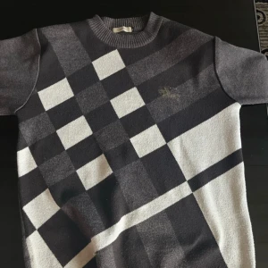 Rutig stickad tröja från Burberry  - Snygg stickad tröja från Burberry London i svart, grått och vitt med klassiskt rutigt mönster. Tröjan har rund halsringning och korta ärmar. Perfekt för dig som gillar stilren design med en twist.