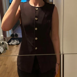 Brun väst med guldknappar - Snygg brun väst med rund hals och sex dekorativa guldknappar framtill. Västen har en figurnära passform och är tillverkad i ett slätt, mjukt material som ger en stilren look. Perfekt att styla över en t-shirt eller skjorta för en trendig vibe.