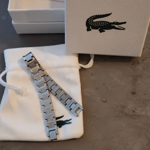 Två armband från Lacoste - Säljer två snygga armband från Lacoste. Ett är i silverfärgad metall med rektangulära länkar och ingraverade krokodiler, det andra är ett tunnare guldarmband med Lacostes ikoniska krokodilberlock. Perfekt för dig som gillar stilrena detaljer och vill visa upp din Lacoste-vibe. Helt nya köpte en till mig och en till min tjej skulle ge present 🎁 men det blev förändringar så nu säljer jag dem för att det har gått 1månad så kan inte skicka tillbaka  Både säljs för 1500kr tillsamman