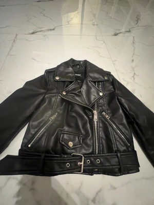 Svart skinnjacka Pull&Bear XS - Snygg svart skinnjacka från Pull&Bear i storlek XS. Jackan har klassisk bikerstil med silvriga dragkedjor, knappar och bälte i midjan. Långärmad och med coola detaljer som snedställd dragkedja och fickor framtill. Perfekt för dig som gillar edgy streetstyle.
