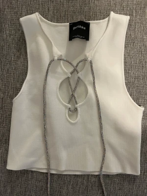 Vit croppad linnetopp från Bershka - Säljer en vit croppad linnetopp från Bershka i storlek M. Toppen har glittriga snörningsdetaljer framtill och är ärmlös med rundad hals. Materialet är mjukt och stretchigt, perfekt för att skapa en cool och trendig look.
