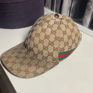 Beige Gucci keps  - Säljer en beige keps från Gucci med klassiskt GG-monogram över hela kepsen. På sidan finns de ikoniska gröna och röda ränderna. Kepsen har böjd skärm och justerbart spänne bak. Materialet är bomullscanvas med detaljer i brunt. Perfekt för dig som gillar lyxiga accessoarer. Söker gärna byte mot svart gucci canvas keps också, hör av er vid funderingar