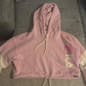 Rosa croppad hoodie från FILA XS - Säljer en ljusrosa croppad hoodie från FILA i storlek XS. Tröjan har huva med snörning, vita detaljer på ärmarna och ett stort FILA-tryck framtill. Perfekt för dig som gillar sportig och chill stil.såljed billigare då den är i använd skick
