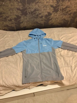 Blå och grå vindjacka från Under Armour storlek S/M - Snygg tvåfärgad vindjacka från Under Armour i ljusblått och grått. Jackan har huva, hel dragkedja framtill och två sidofickor. Perfekt för dig som gillar sportig stil och vill ha något lätt och funktionellt. Logga på bröstet för extra vibe.