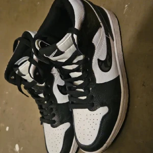 Nike Jordan 1 svartvita sneakers - Svartvita Nike Jordan 1 sneakers med högt skaft och klassisk snörning. Skorna har en rund tå, platt sula och är tillverkade i skinn med perforerade detaljer på tån. Swoosh-loggan syns tydligt på sidan. Perfekt för dig som gillar streetwear och sportig stil.