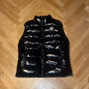 Svart dunväst från Moncler - Snygg svart dunväst från Moncler med glansig finish och klassisk logga på bröstet. Västen har ståkrage, två fickor med dragkedja och quiltad design. Insidan har ett coolt serietidningsmönster på tvättlappen. Perfekt för lager-på-lager och streetstyle.