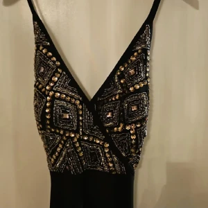 Svart långklänning med paljetter nypris 3500 - Svart långklänning med smala axelband och djup v-ringning. Överdelen är dekorerad med glittriga paljetter och geometriska mönster i guld och silver, vilket ger en lyxig och festlig vibe. Kjolen är enkel och faller rakt ner. Perfekt för dig som vill sticka ut.