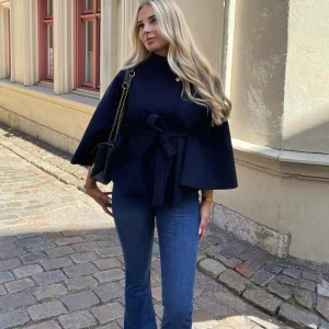 Mörkblå bolerotopp från Missay - Snygg mörkblå bolerotopp från Missay med hög krage, guldfärgad knapp och knytband i midjan. Toppens cape-liknande form ger en cool och elegant vibe. Perfekt att styla med jeans eller kjol för en trendig look.