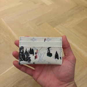 Vit plånbok med vintermotiv - Snygg vit plånbok i skinn med vinterinspirerat tryck av skidåkare, lift och snötäckta granar. Perfekt för kort och sedlar, med flera fack på båda sidor. Unik och lekfull design som sticker ut i mängden.