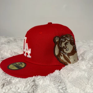 Röd LA keps New Era 59FIFTY 7 1/4 - Säljer en röd LA Dodgers keps från New Era 59FIFTY med broderad vit LA-logga framtill och en stor broderad ängel på sidan. Baksidan har texten 'Ion Stan' i guldfärgad brodyr. Platt skärm och klassisk passform.
