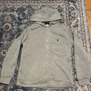 Grå hoodie från Polo Ralph Lauren - Säljer en grå hoodie från Polo Ralph Lauren med dragkedja och huva. Tröjan har långa ärmar, två fickor framtill och den klassiska Polo-loggan broderad på bröstet. Perfekt för chill dagar och enkel att matcha med jeans eller joggers.
