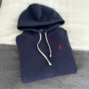 Marinblå hoodie från Polo Ralph Lauren - Snygg marinblå hoodie från Polo Ralph Lauren med vit snörning och klassisk röd logobrodyr på bröstet. Tröjan har huva, långärmad design och en stor magficka framtill. Perfekt för chill dagar och enkel att matcha med jeans eller joggers. Storlek xs