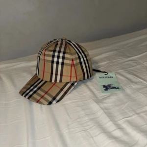 Burberry rutig keps med logga - Snygg Burberry keps i klassiskt beige, svart, vit och röd rutigt mönster. Framtill med böjd skärm och baktill justerbar snapback. Märkt med Burberry London England på baksidan. Tillverkad i bomull med svart insida. Perfekt för dig som vill ha en ikonisk streetstil.