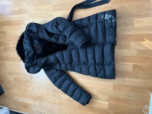 Svart dunjacka med fuskpäls och bälte - Snygg svart dunjacka från Red Label med fluffig fuskpäls på huvan och praktiskt bälte i midjan. Jackan har långa ärmar, två dragkedjefickor och quiltad design som ger en cool look. Perfekt för kalla dagar och riktigt varm.