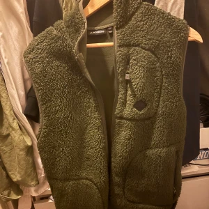 Grön fleeceväst från J.Lindeberg - Mysig grön fleeceväst från J.Lindeberg i storlek M. Västen har en fluffig, teddy-liknande yta, praktisk bröstficka med dragkedja och hel dragkedja framtill. Perfekt för lager-på-lager och chill stil. Snygg och enkel att matcha med hoodies eller t-shirts. Pris kan diskuteras.