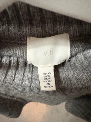Cropped grå stickad tröja från H&M storlek XS - Säljer en grå croppad tröja från H&M med ett snyggt kabelstickat mönster. Tröjan är i storlek XS. Väl använd och lite nopprig på ryggen, men inget som syns. 